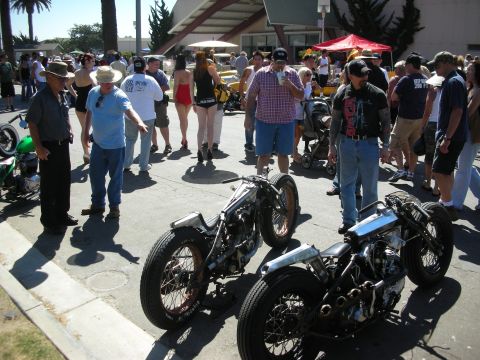 1998 Ford Crown Victoria - Spectators in hats and vests gather around two low bare-metal hardtail bobbers parked on a sunny outdoor show street.