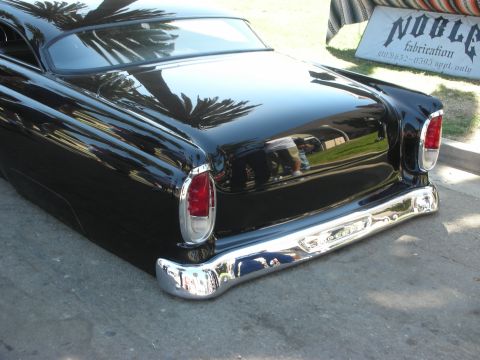 1998 Ford Crown Victoria - Rear quarter of a glossy black chopped custom with frenched taillight and chrome bumper, near a NOBLE fabrication banner.