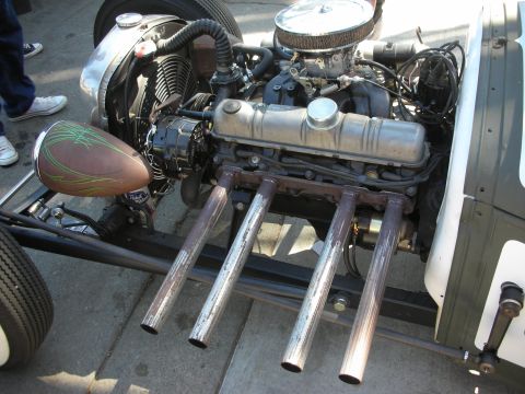 1998 Ford Crown Victoria - Close-up of a rat rod's exposed engine, carburetor and four upright open chrome header pipes beside a green pinstriped tank.