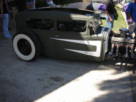 1998 Ford Crown Victoria - heavily chopped and channeled olive-green rat rod sedan with whitewall tires sits low at a curbside car show with its engine exposed.