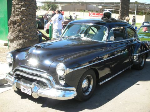 1998 Ford Crown Victoria - black Oldsmobile fastback sedan with chrome trim and a yellow license plate sits under palm trees at an outdoor car show.