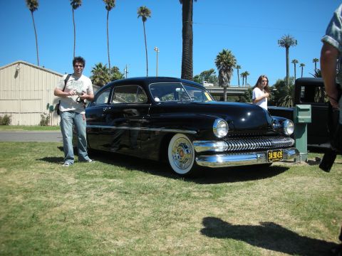 1998 Ford Crown Victoria - chopped black Mercury lead-sled custom coupe sits on grass under palm trees, wearing wide whitewalls and a yellow license plate.