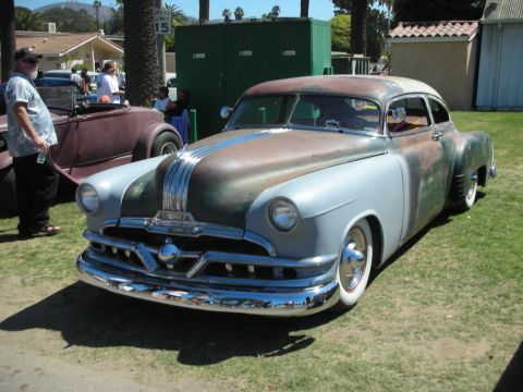 1998 Ford Crown Victoria - chopped Pontiac lead-sled custom in faded silver with rust patina and a chrome grille sits low on grass at an outdoor car show.