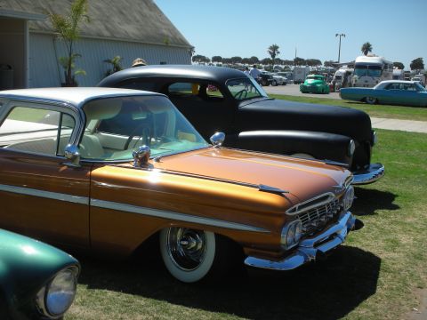 1998 Ford Crown Victoria - copper-and-white 1959 Chevrolet with cat's-eye taillights sits beside a flat-black chopped custom on a grass car-show field by the coast.