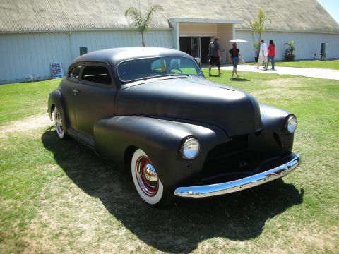1998 Ford Crown Victoria - flat-black chopped 1940s coupe custom with red-trim wheels and whitewall tires sits on a grass field at a sunny car show.