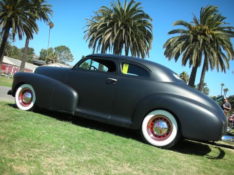 1998 Ford Crown Victoria - side profile of a flat-black chopped fastback coupe with red-painted wheels and whitewalls sits on grass under tall palm trees.