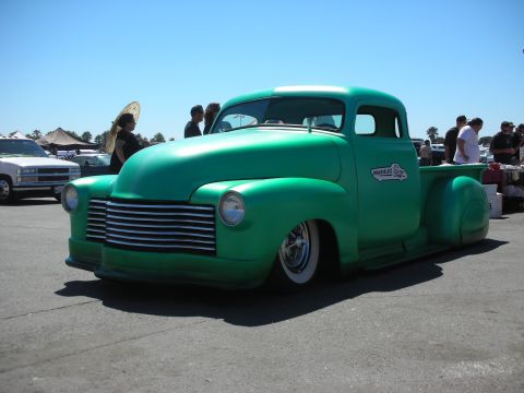 1998 Ford Crown Victoria - bright-green chopped late-1940s Chevrolet pickup sits slammed low with chrome grille and whitewalls on a paved car-show lot.