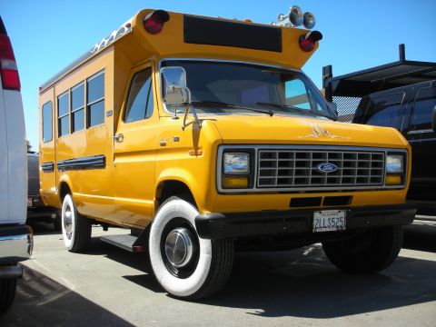 1998 Ford Crown Victoria - yellow Ford short school bus with flame graphics, roof lights, whitewalls, and a chrome grille sits in a car-show parking lot.