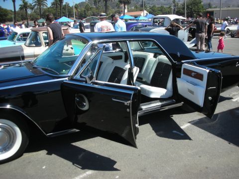 1998 Ford Crown Victoria - black Lincoln Continental sedan sits with its suicide rear door open, revealing a black-and-white interior, at a crowded coastal car show.