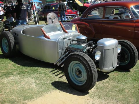 1998 Ford Crown Victoria - bare aluminum-bodied roadster hot rod with a green V8 engine and steel wheels displayed on grass at a car show.