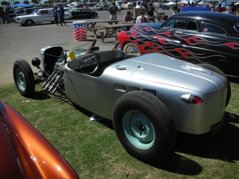 1998 Ford Crown Victoria - Rear three-quarter view of a silver boattail roadster hot rod with green steel wheels beside a flamed custom at a car show.