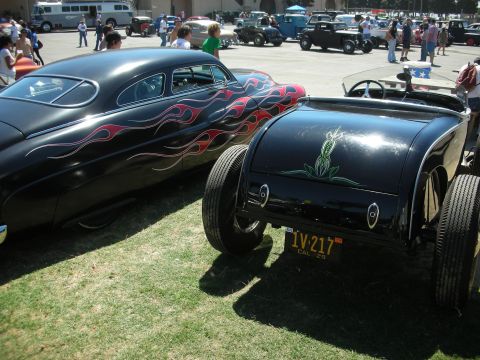1998 Ford Crown Victoria - black hot rod roadster with pinstriped decklid and California plate parked next to a red-flamed custom coupe on grass.