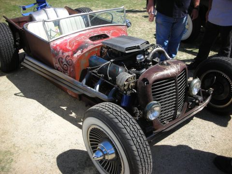 1998 Ford Crown Victoria - rat rod roadster with red swirl-painted body, patina grille shell, exposed V8 and whitewall tires at an outdoor show.