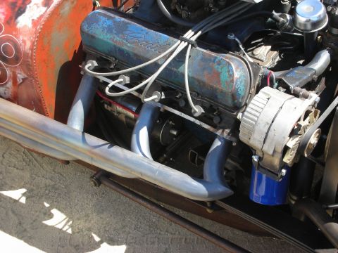 1998 Ford Crown Victoria - Close-up of a patina blue V8 engine with cast valve cover, alternator and tubular headers in a rat rod.