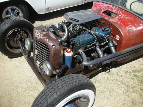 1998 Ford Crown Victoria - Front engine bay of a rat rod showing patina grille shell, blue V8, headers and red swirl-painted body at a car show.
