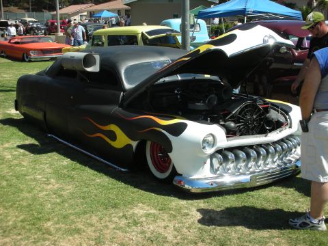 1998 Ford Crown Victoria - black chopped lead-sled custom with yellow flames, open hood, chrome toothy grille and red wheels at a car show.