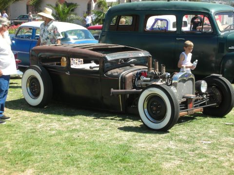 1998 Ford Crown Victoria - brown patina chopped sedan rat rod with exposed engine, whitewall tires and a boy walking past at an outdoor show.