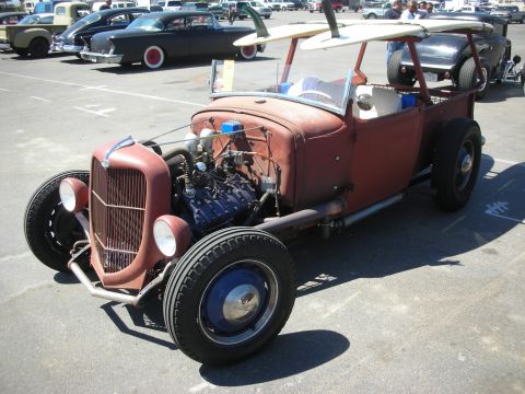 1998 Ford Crown Victoria - brown patina roadster pickup rat rod carrying a surfboard, with exposed V8 and blue wheels at a parking-lot car show.