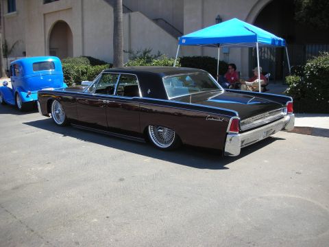 1998 Ford Crown Victoria - brown lowered 1960s Lincoln Continental four-door sedan laid out on the pavement near a blue truck and canopy.