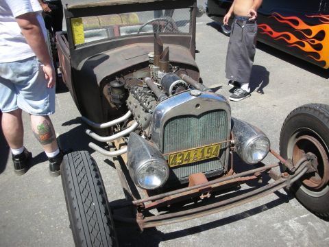 1998 Ford Crown Victoria - rusty bare-metal rat rod with an exposed inline engine and chrome headlights sits front-on among onlookers at a car show.