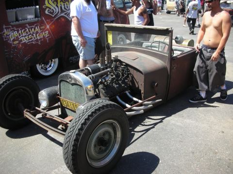 1998 Ford Crown Victoria - rust-patina rat rod roadster with an exposed engine parked beside a graffiti-painted van as people stand around at a show.