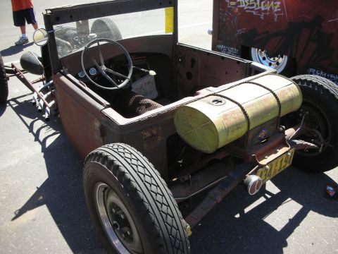 1998 Ford Crown Victoria - rear of a rusty open rat rod roadster reveals a cylindrical barrel fuel tank, steering wheel and bench seat in the open cockpit.