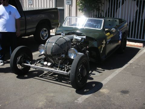 1998 Ford Crown Victoria - olive-green military-themed hot rod roadster with a screened nose and exposed front axle parked beside a pickup at a show.