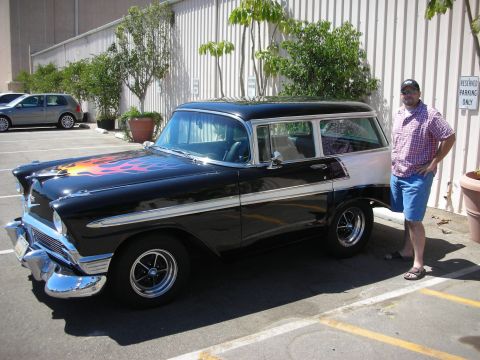 1998 Ford Crown Victoria - man in a plaid shirt stands beside a black 1950s two-door station wagon with hood flames and chrome trim in a parking lot.