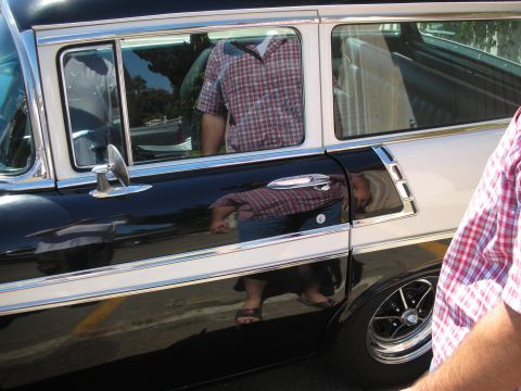 1998 Ford Crown Victoria - Close-up of a black station wagon's rear door and chrome trim, with a man reflected in the glossy paint and bystander at right.