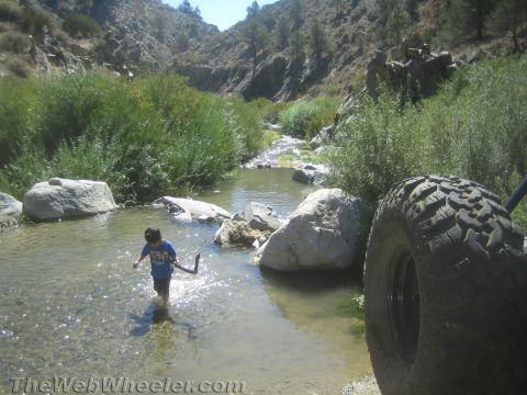1987 Suzuki Samurai - A child plays in a rocky stream surrounded by lush greenery and hills under a clear sky.