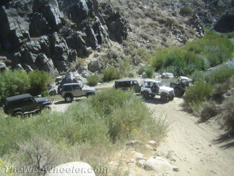 1987 Suzuki Samurai - Several off-road vehicles are gathered on a dirt path next to a stream with a rocky cliff face in the background.