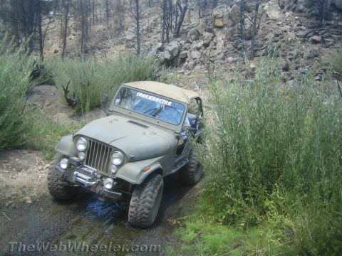 1987 Suzuki Samurai - A Jeep CJ-7 navigates a muddy, off-road trail surrounded by vegetation and burnt trees.