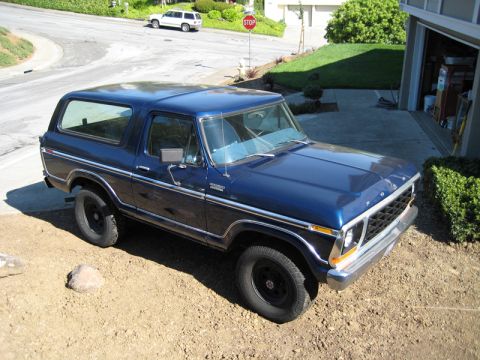 1978 Ford Bronco parked in a driveway, blue paint, chrome trim, black wheels. White SUV visible on street