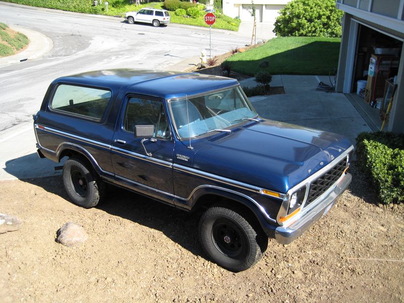1978 Ford Bronco parked in a driveway, blue paint, chrome trim, black wheels. White SUV visible on street