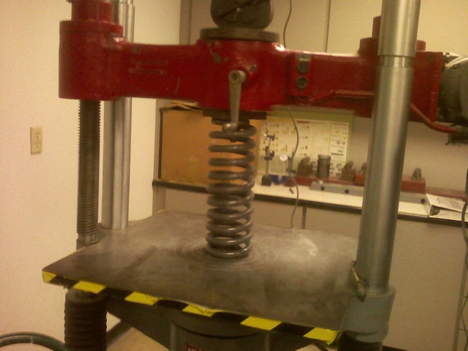 Coil spring standing on the platen of a red hydraulic press in a shop