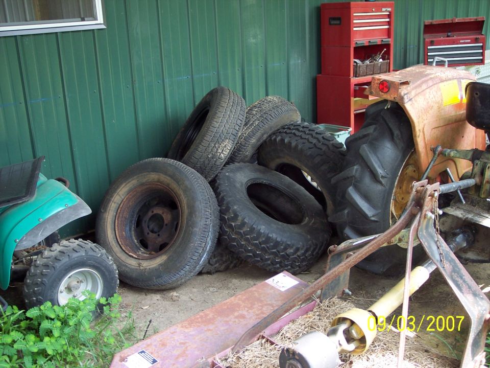 Pile of tires leaning against a green wall, with parts of a tractor and a small green ATV visible