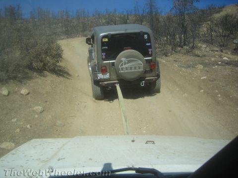 1987 Suzuki Samurai - A Jeep Wrangler Sahara is being towed up a dirt trail by another vehicle, with burnt trees in the background.