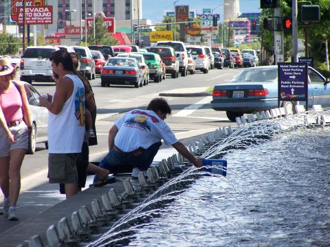 1990 Ford Bronco - A man adds something to a fountain while traffic moves along a street lined with businesses and pedestrians.