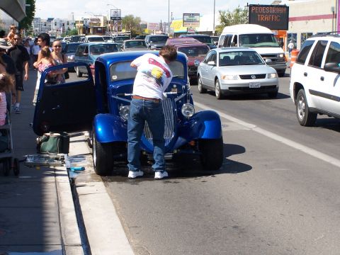 1990 Ford Bronco - A blue hot rod is parked on the street with its door open as a man works on it, surrounded by traffic.