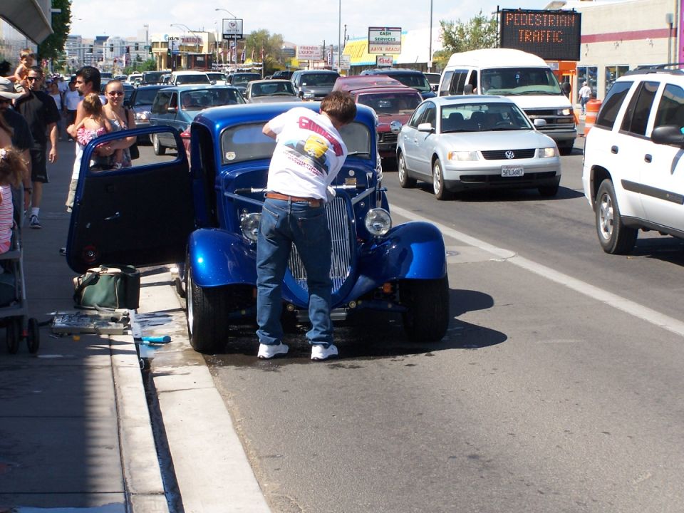 Blue hot rod with open door, man working on engine, traffic behind