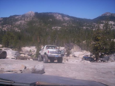 1989 Ford Bronco - A white SUV is parked on a dirt road with rocky terrain and trees in the background.