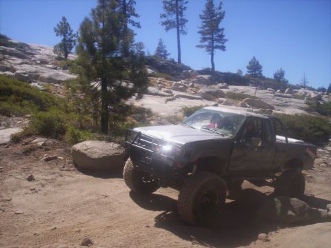 1989 Ford Bronco - A heavily modified pickup truck navigates a , rocky off-road path under a clear blue sky.