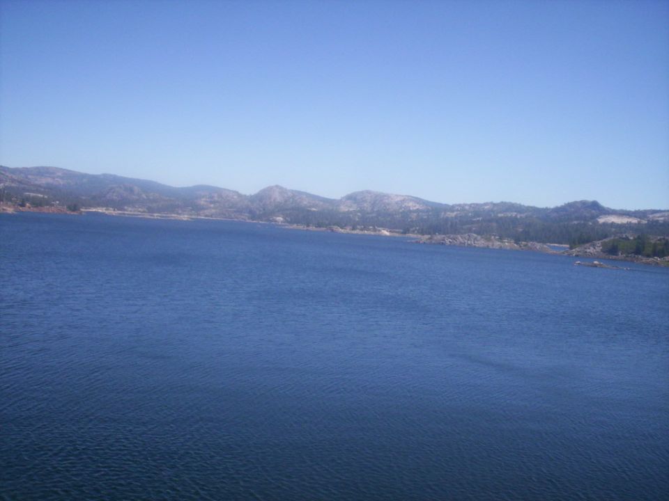 Wide view of a blue lake with tree-covered mountains in the background under a clear blue sky