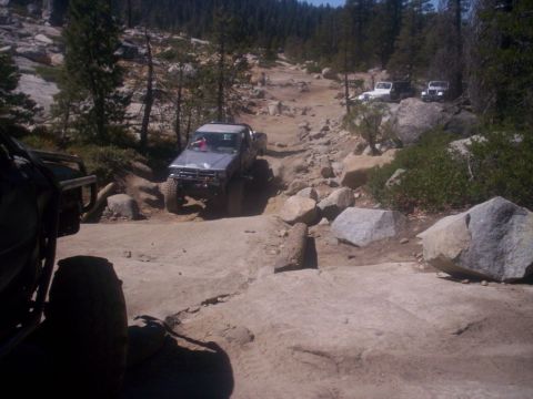 1989 Ford Bronco - Several off-road vehicles navigate a challenging, rocky terrain under a clear sky with trees in the background.