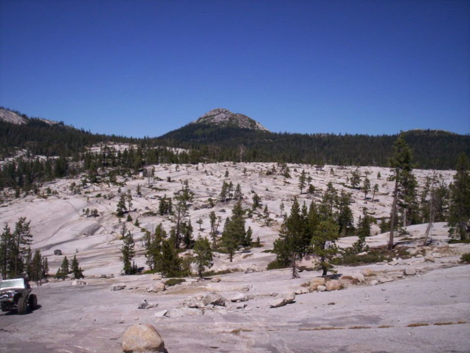 Front view of a dark-colored Jeep Wrangler parked on a rocky, mountainous terrain with scattered trees