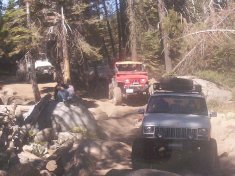 1989 Ford Bronco - Several off-road vehicles navigate a wooded trail with rocky terrain.