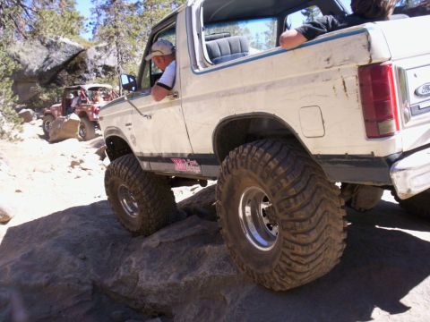 1989 Ford Bronco - A white 1989 Ford Bronco, modified with large off-road tires, tackles a challenging rocky trail with another vehicle visible behind it.