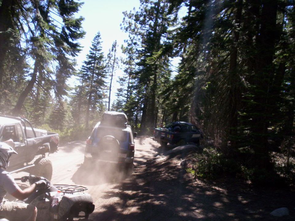 Dusty dirt road with three off-road vehicles, including a blue SUV and a pickup truck, driving through a forest