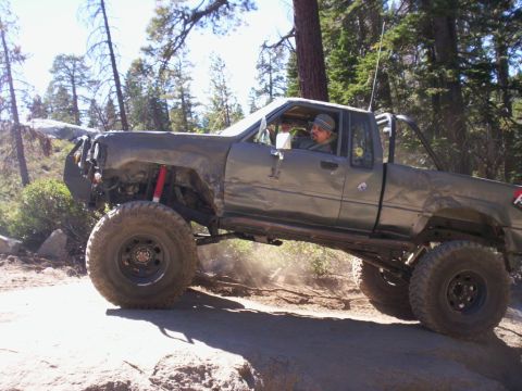 1989 Ford Bronco - A heavily modified pickup truck navigates a challenging off-road trail, kicking up dust as it goes.
