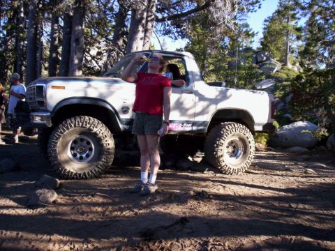 1989 Ford Bronco - A person takes a break next to a lifted Ford Bronco on a dirt road surrounded by trees.